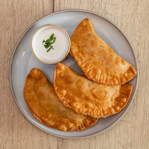 Three golden-brown fried empanadas and a small bowl of white dipping sauce with a cilantro garnish.