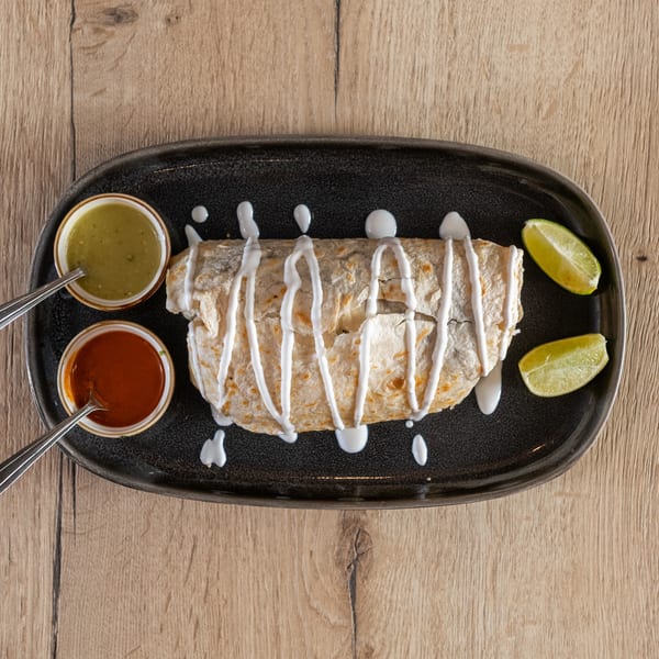A burrito drizzled with white sauce, served with two lime wedges and two small bowls of salsa.