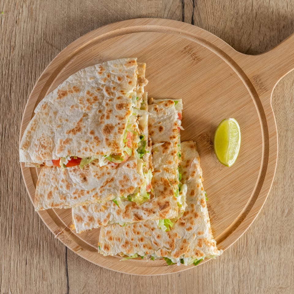 Four quesadilla slices filled with lettuce, tomato, and cheese, served with a lime wedge on a wooden board.